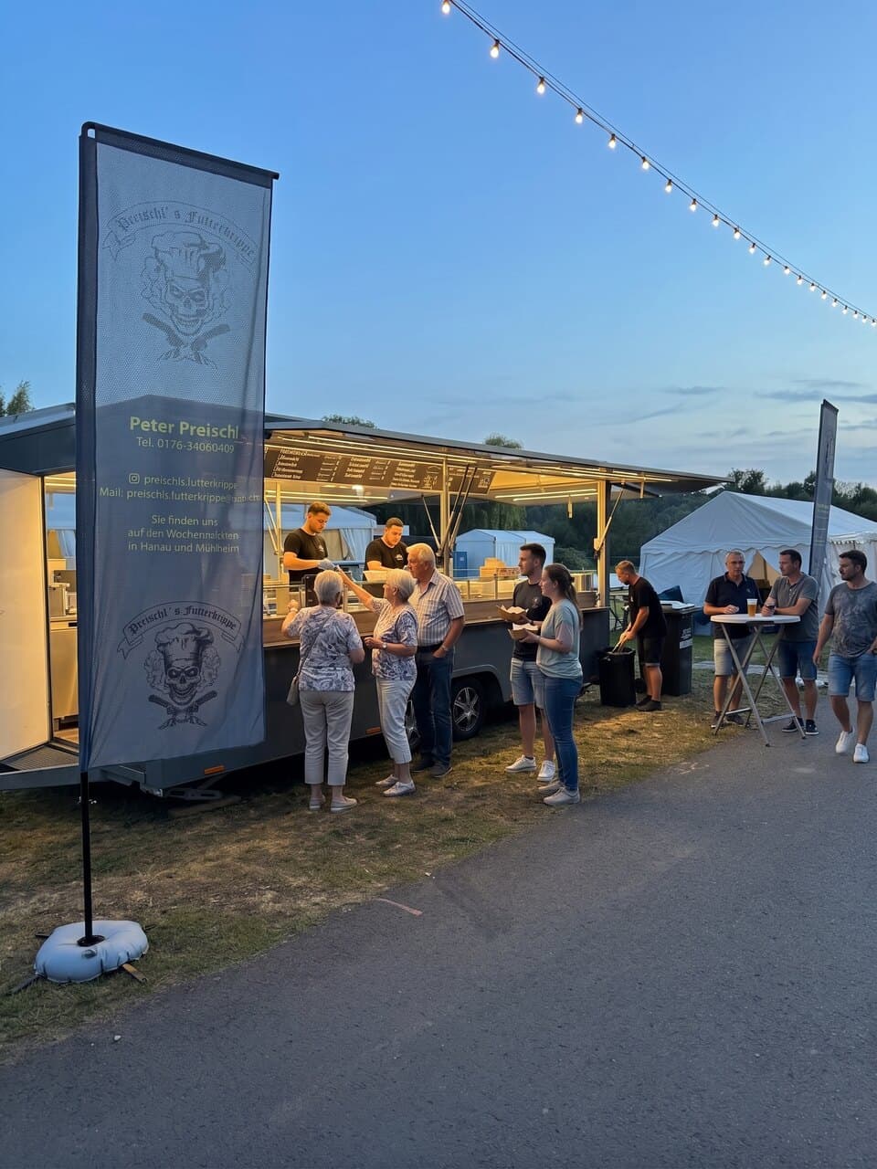 Foodtruck in der Abenddämmerung mit Lichterkette und Banner, Gäste werden bedient