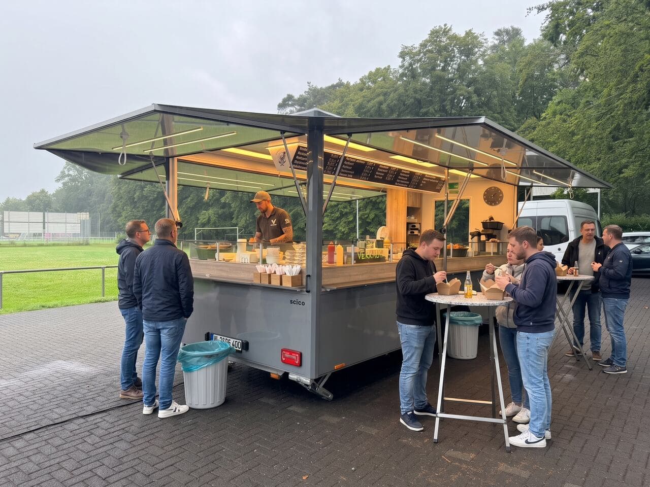 Foodtruck bei einem Vereinsfest, Gäste essen am Stehtisch unter bedecktem Himmel